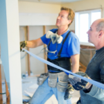 One person wearing a blue shirt holding a tape measure, while another man holds it at the other end, measuring a door way of a property they are renovating.