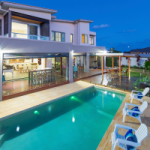 An unhosted Airbnb property, with a pool, lit up with with pool lights at the rear of the house. Pool chairs line the side of the pool. Short term rental properties like this one will have the number of days it can be rented out capped in Byron Bay to encourage long term rentals in the area.