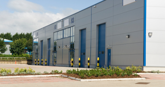 A row of grey warehouses with blue doors and yellow and black safety bollards. Warehouses a popular choice for new commercial property investors because the entry point is low and the building is very simple to manage.