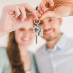 A young couple hold the keys out to their first home. They have used the First Home Owners Grant to purchase the property. This means they must live in the property for 12 months before renting it out. This means that the Assets will be considered second hand on rental.